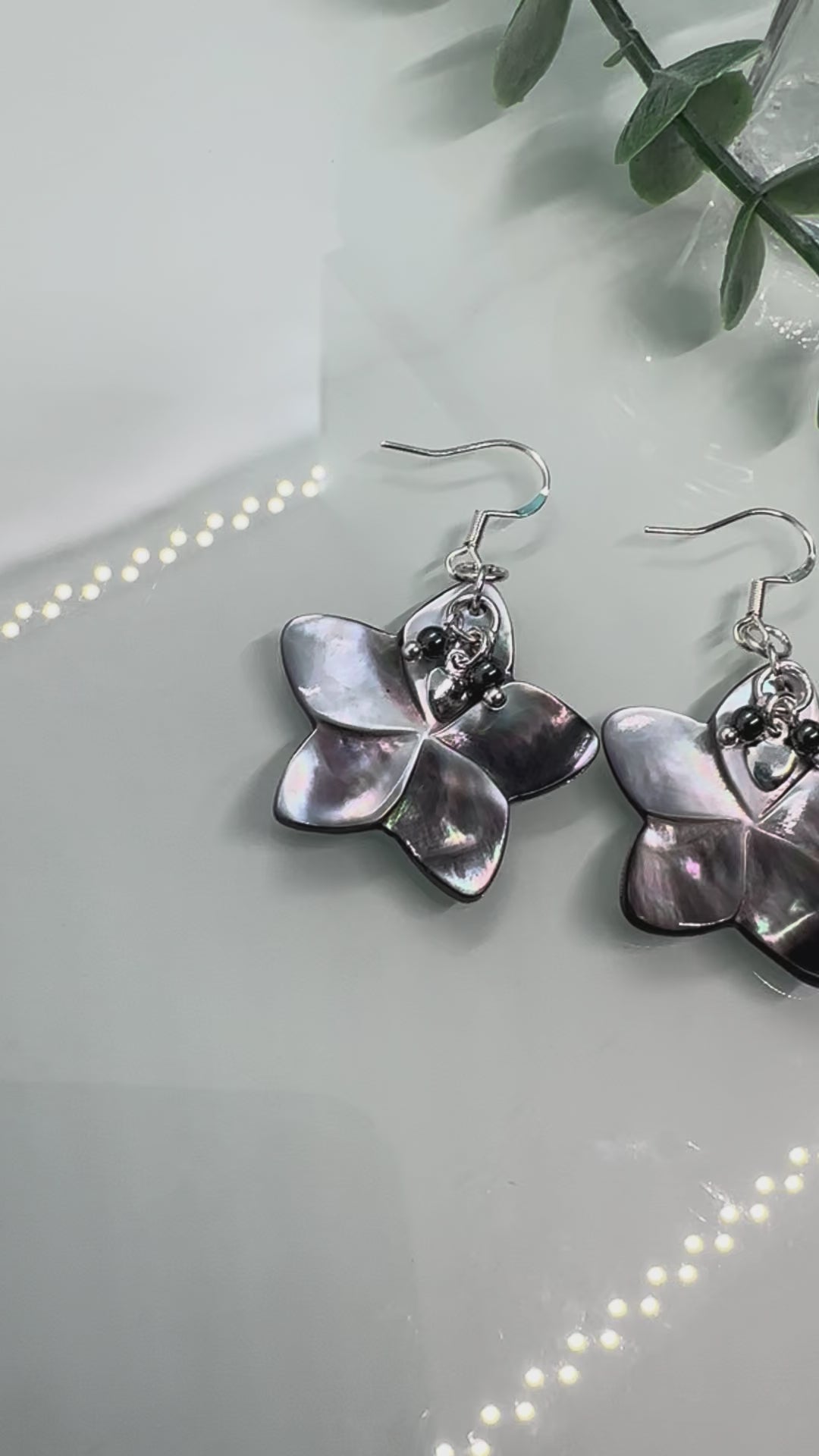 Video showing Abalone shell carved flower shapes with iridescent grey shell petals, and with small Hematite and silver hearts layered on top, drop earrings with sterling silver components